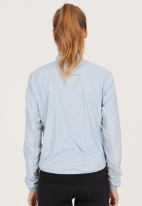 Hellblaues Sportjacke mit glatter Textur, Netzdetails an den Ärmeln, elastischen Bündchen und einem schwarzen Saum. Auf der Rückseite befindet sich ein Schallwellen-Design.