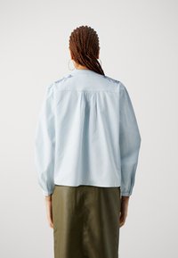 Blusa a righe azzurra chiara con maniche lunghe, vestibilità morbida e ricamo floreale vicino al collo; abbinata a una gonna di pelle verde.