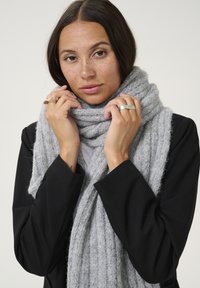 Écharpe grise en tricot avec une texture côtelée, portée lâchement autour du cou, associée à un blazer noir. Douceur et chaleur remarquables.