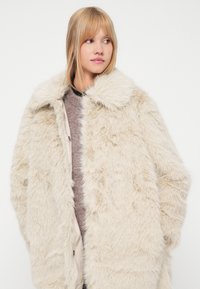 Krēmveida mākslīgā kažokāda ar plašu apkakli, priekšējiem pogām un mīkstu tekstūru. Valkā virs pelēka adīta krekla.