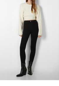 Vrouw in een witte cropped trui, hoge zwarte skinny broek en zwarte enkelhoge haklaarzen, staande tegen een witte achtergrond.