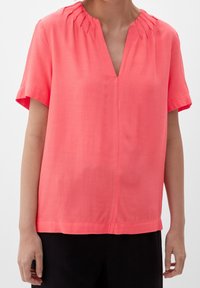 Blouse corail à manches courtes, encolure en V et détails plissés aux épaules. Tissu lisse et coupe décontractée. Pantalon noir visible en bas.