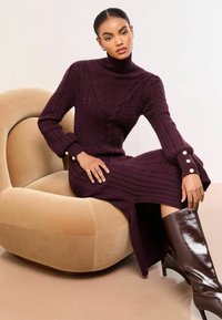 Vīnogu krāsas adīts džemperkleita ar kabeļu rakstu, pieguļošu augstu apkakli un dekoratīvām pogu detaļām piedurknēs, kombinēta ar spīdīgām brūnām ceļgalu augstām zābakiem.