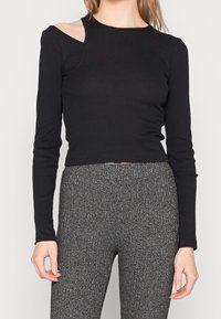 Schwarzes Langarmoberteil mit gerippter Textur und asymmetrischem Schulter-Ausschnitt-Design. Kombiniert mit grauen hochtaillierten Hosen mit Struktur.