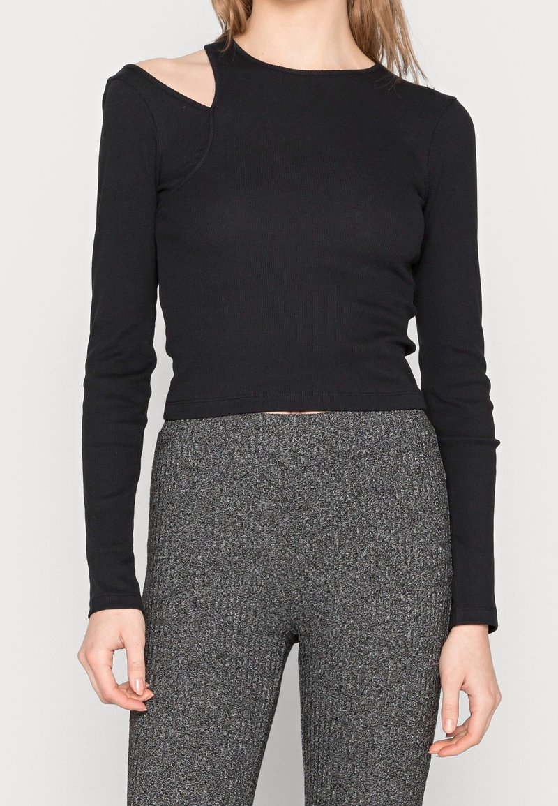 Schwarzes Langarmoberteil mit gerippter Textur und asymmetrischem Schulter-Ausschnitt-Design. Kombiniert mit grauen hochtaillierten Hosen mit Struktur.