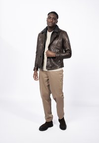 Chaqueta de cuero marrón con un collar negro, suéter ligero debajo, pantalones beige y botas oscuras. Textura suave y diseño ajustado.