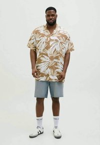 Mand iført en lysebrun og hvid kortærmet skjorte med bladmønster, lyse denimshorts, hvide sokker og hvide sneakers, står foran en ensfarvet baggrund.