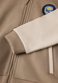 Beige zip-up jakke med ribbet, cremefarvet manchet og et cirkulært mærke med et landskabsdesign. Tekstureret stof med glatte accenter.