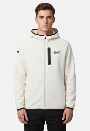 Giovane uomo che indossa una felpa bianca con zip con logo EA7 Emporio Armani e rifiniture nere, in piedi davanti a uno sfondo semplice.
