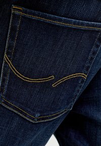 Temno modri žepi na hrbtni strani kavbojk iz jeansa z oranžnimi šivi in ukrivljenim okrasnim šivom na tkanini.