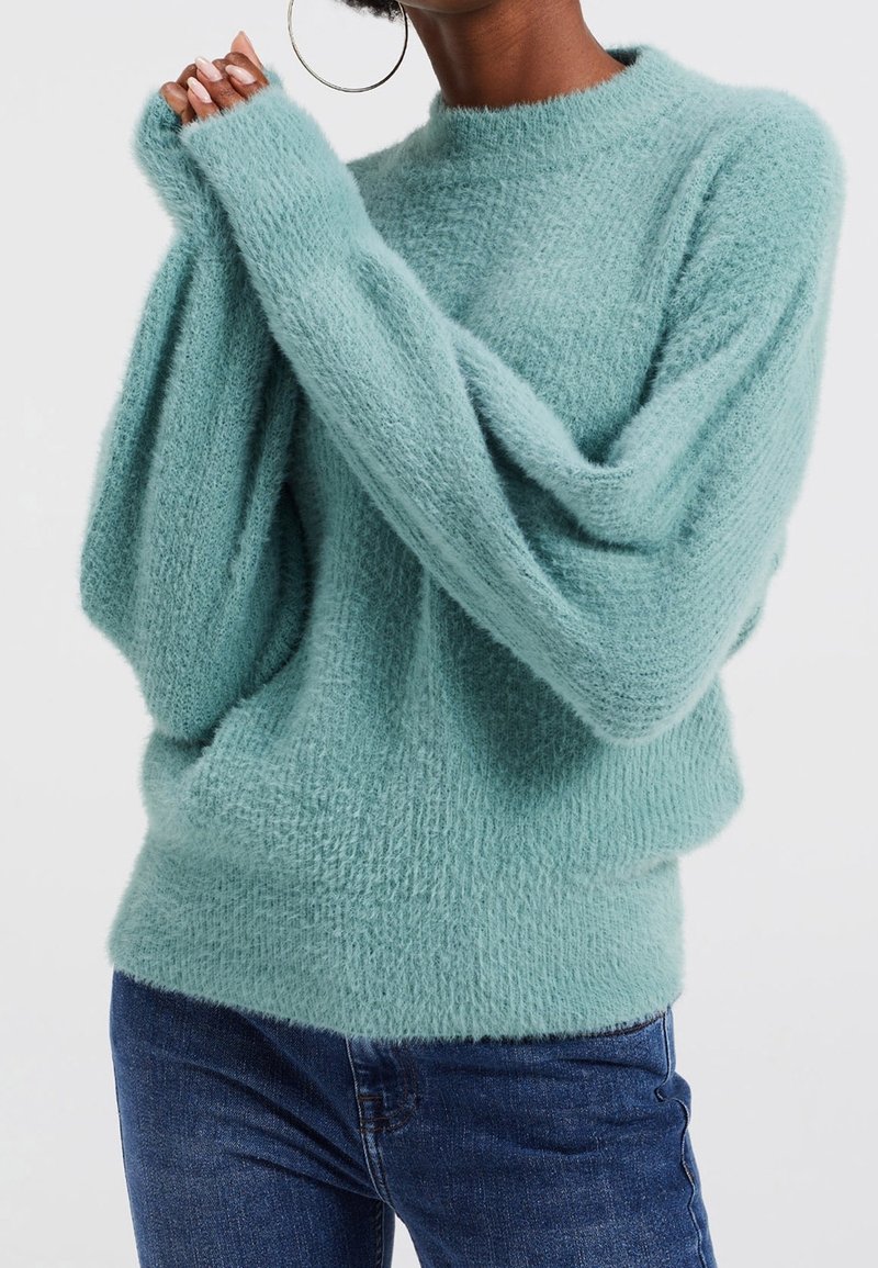 Personne portant un pull doux bleu clair et un jean bleu foncé, les mains levées près du visage, avec de grandes boucles d'oreilles en forme d'anneau visibles.
