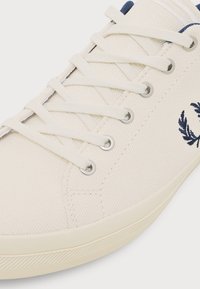 Gros plan d'une basket en toile blanche avec des lacets blancs, des œillets argentés et un logo de couronne de laurier brodé en bleu marine sur le côté.