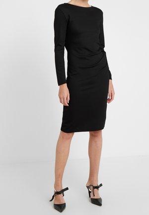 Kobieta ubrana w dopasowaną czarną sukienkę z długim rękawem sięgającą do kolan z marszczeniem po bokach oraz czarne szpilki z kokardkami przy kostkach.