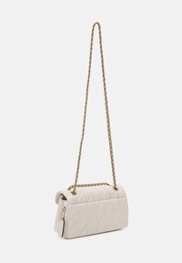 Kremowa pikowana torebka na ramię z złotym łańcuszkiem i zwisającą prostokątną metką na białym tle.