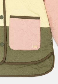 Quiltet jakke med gule, pink og grønne sektioner. Har en pink lomme, subtil blomsterbroderi og mærketag i nederste hjørne.