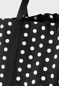 Μαύρη τσάντα tote από ύφασμα με λευκά πουά και μαύρες υφασμένες λαβές, δείχνοντας μέρος του εσωτερικού με μια μικρή ετικέτα.