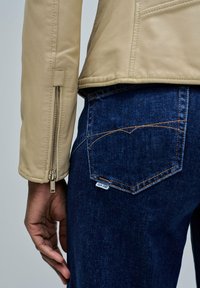 Main reposant près de la poche d'un jean en denim bleu avec des surpiqûres orange, portant une veste en cuir beige avec des détails de fermeture éclair sur les manches.