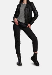 Czarna skórzana kurtka motocyklowa na szarym T-shircie, w połączeniu z czarnymi przylegającymi dżinsami i masywnymi czarnymi botkami za kostkę. Modelka pozuje z jedną ręką na biodrze.