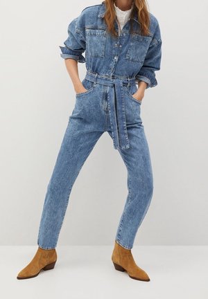 Jeans-Overall in Hellblau mit durchgehender Knopfleiste, tailliertem Gürtel, zwei Brusttaschen und zulaufenden Beinen. Abgerundet mit cognacfarbenen Stiefeletten.