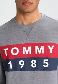 Sudadera gris con llamativas rayas horizontales en rojo y marino. El texto dice "TOMMY JEANS 1985." Cuello redondo y textura suave.