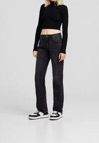 Svart räfflad crop-top, högmidjade mörka jeans och svarta och vita sneakers med kontrasterande sula och snören, på en ljusgrå bakgrund.