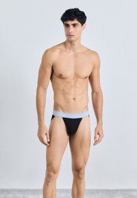 Crna tanga donji veš s svijetloplavim elastičnim pojasom na kojem se nalazi logo "Calvin Klein". Jednostavan dizajn, glatka tekstura i minimalna pokrivenost.