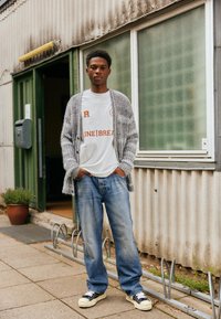 Cardigan grigio lavorato a maglia, comodo, indossato su una t-shirt bianca con testo stampato, abbinato a jeans blu slavati e sneakers nere con suole bianche.