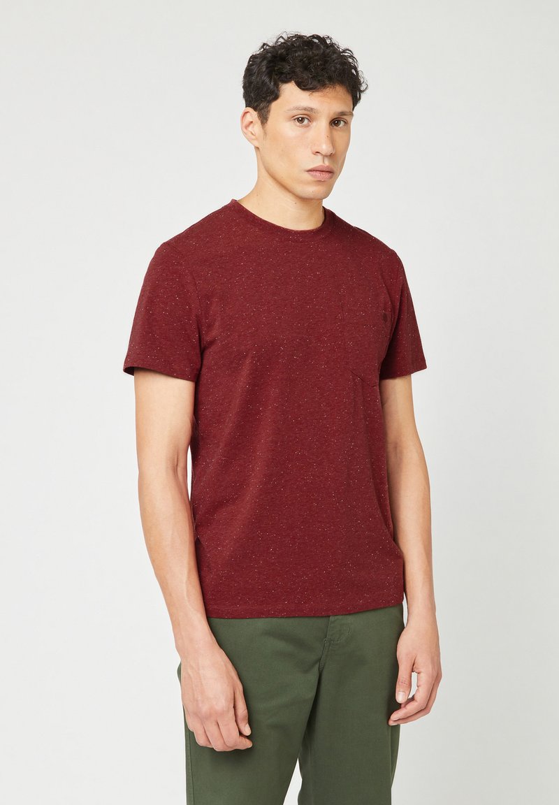 Jonge man met kort, krullend donker haar, gekleed in een kastanjebruingevlekt shirt met korte mouwen en olijfgroene broek, staand tegen een effen achtergrond.