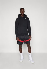 Čierna mikina s kapucňou so šnúrkami a klokaním vreckom, s zeleným logom NBA; doplnená o čierne šortky s červenými pruhmi; biele tenisky; jednoduchý, športový štýl.