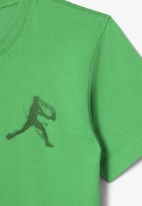 Vihreä puuvillapaita, jossa on tummanvihreä siluetti graafinen kuva liikkuvasta pelaajasta, pyöreä pääntie ja lyhyet hihat.