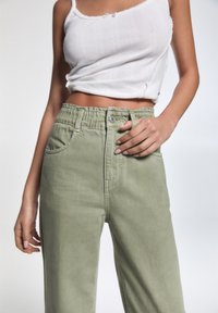 Pantalones de mezclilla verde con cintura elástica, bolsillos frontales y un botón plateado. Combinados con una camiseta corta de tirantes acanalada blanca.