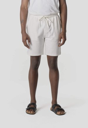 Persona vistiendo pantalones cortos blancos con textura y cordón, camisa blanca y sandalias negras de doble tira, de pie sobre un fondo liso y claro.