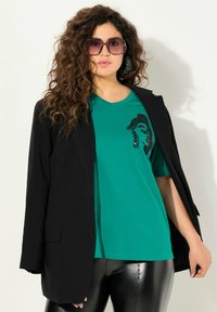 Blazer nero sopra una t-shirt a v blu petrolio con una grafica di un volto femminile. Il modello indossa occhiali da sole grandi e leggings neri lucidi.