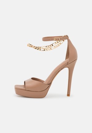 Beige High-Heel-Sandale mit Plateau, Knöchelriemen und zwei goldenen Kettendetails auf weißem Hintergrund.