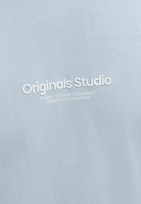 Šviesiai mėlynas medvilninis marškinėliai su iškilniu balto teksto užrašu: "Originals Studio," papildomu tekstu žemiau. Lygus paviršius, kasdieniškas dizainas.