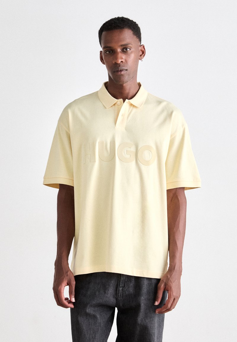 Gelbes Polohemd mit einem Kragen mit drei Knöpfen und geprägtem "HUGO"-Logo auf der Vorderseite, kombiniert mit dunklen Jeans. Kurzärmelig und lockere Passform.