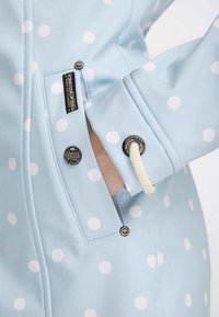 Main dans la poche d'une veste bleu clair à pois avec des boutons métalliques de marque et une boucle de cordon blanche au poignet.