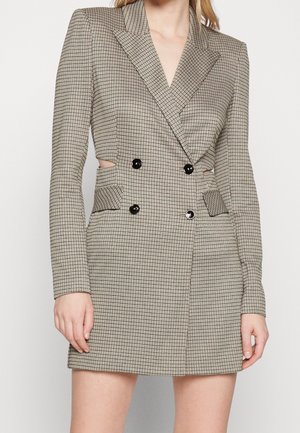 Femme portant une robe blazer à carreaux beige croisée, manches longues et boutons noirs, avec de petites découpes latérales à la taille.
