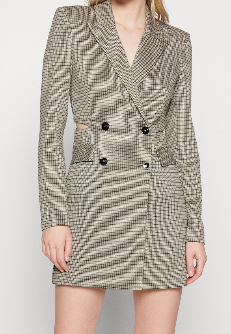 Femme portant une robe blazer à carreaux beige croisée, manches longues et boutons noirs, avec de petites découpes latérales à la taille.