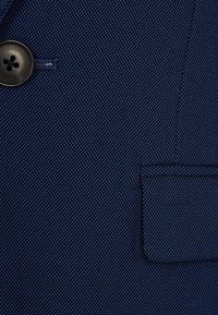 Ciemnoniebieska tekstura materiału z drobnym wzorem kropek, z czarnym guzikiem na cztery dziurki oraz prostokątną klapą kieszeni z materiału.