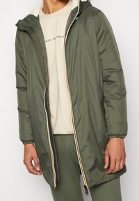 Giacca imbottita verde con chiusura frontale con cerniera, dotata di cappuccio foderato in pelliccia bianca e strisce colorate lungo la cerniera.