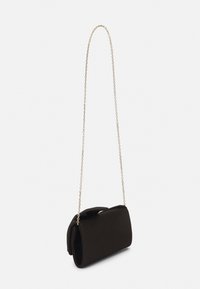 Piccola borsa a tracolla rettangolare nera con chiusura a patta, appesa a una sottile tracolla in oro su uno sfondo semplice.