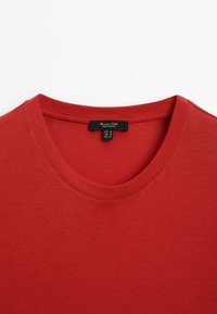 T-shirt en coton rouge avec un col rond, présentant une texture lisse et une étiquette affichant les informations de taille pour plusieurs régions.