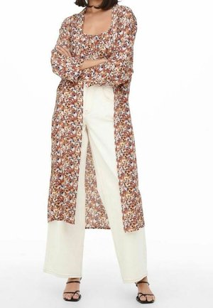 Femme portant un pantalon blanc à jambes larges avec un long cardigan fleuri assorti et un haut, debout, les bras croisés et les pieds en sandales noires.