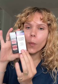 Une personne avec des cheveux blonds bouclés tient et présente un produit de baume à lèvres emballé étiqueté "Baume à Lèvres Repulpant aux Tripeptides."