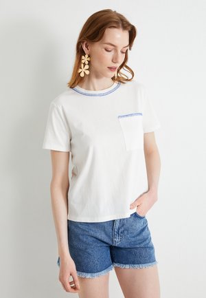T-shirt bianco di cotone con colletto e tasca ricamati in blu, abbinato a pantaloni shorts in denim con bordi sfrangiati.