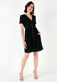 Abito nero a maniche corte con scollatura a V, chiusura frontale con bottoni e gonna arricciata. Caratterizzato da tasche laterali e una texture di tessuto liscia.