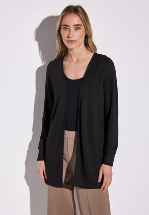 Cardigan long noir avec une ouverture sur le devant, fabriqué en matériau doux. Il possède des manches longues et une coupe décontractée, associé à un haut noir et un pantalon beige.