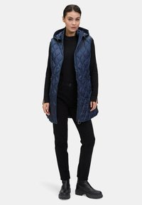 Námornícky modrá prošívaná péřová vesta s kapucí, se zipem vpředu a bočními kapsami, v kombinaci s černými kalhotami a kotníkovými botami.