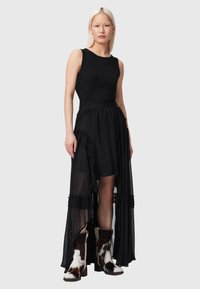 Γυναίκα με μακριά ξανθά μαλλιά, φορώντας αμάνικο μαύρο φόρεμα με διαφανείς φρατζαριστές λεπτομέρειες και μπότες μέχρι τη μέση της γάμπας με μοτίβο αγελάδας, στέκεται μπροστά σε γκρι φόντο.